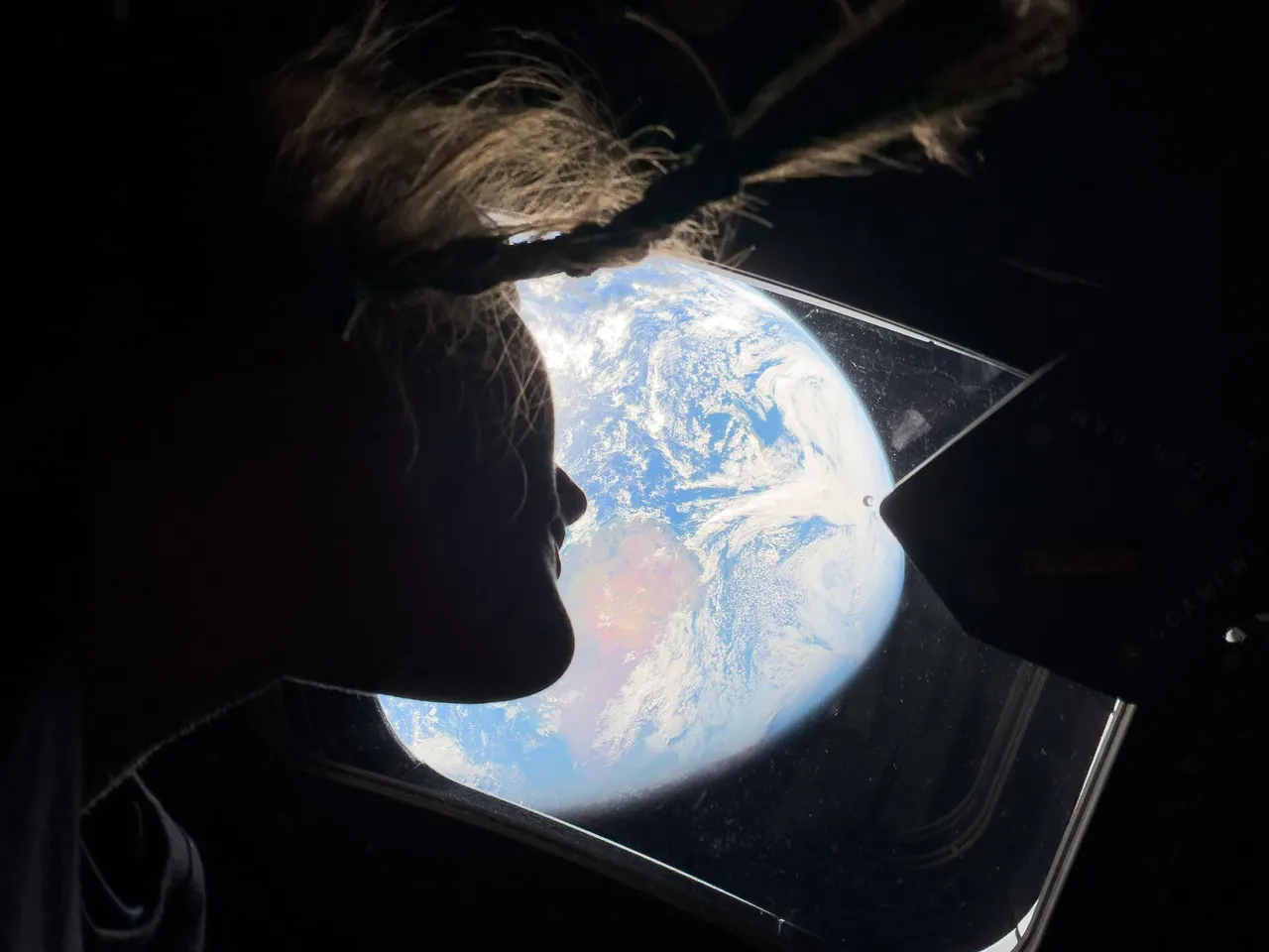 Silhouet van een persoon met gevlochten haar die door het raam van een ruimtecapsule naar de aarde kijkt.